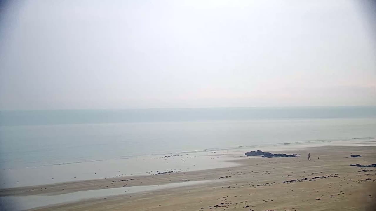 A hazy, calm ocean meets a wide, sandy beach where a lone person walks.
