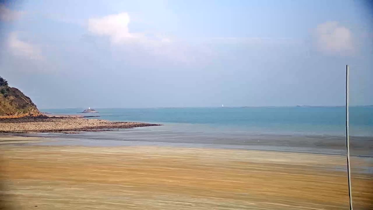 A mostly sunny day shows a wide sandy beach extending to a calm teal ocean, with a small, rocky headland and distant island visible.