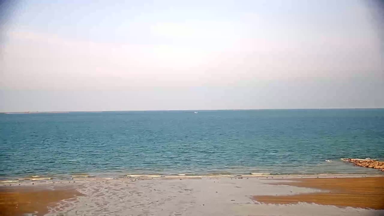 A calm, light-blue ocean meets a wide, sandy beach under a mostly cloudy sky.