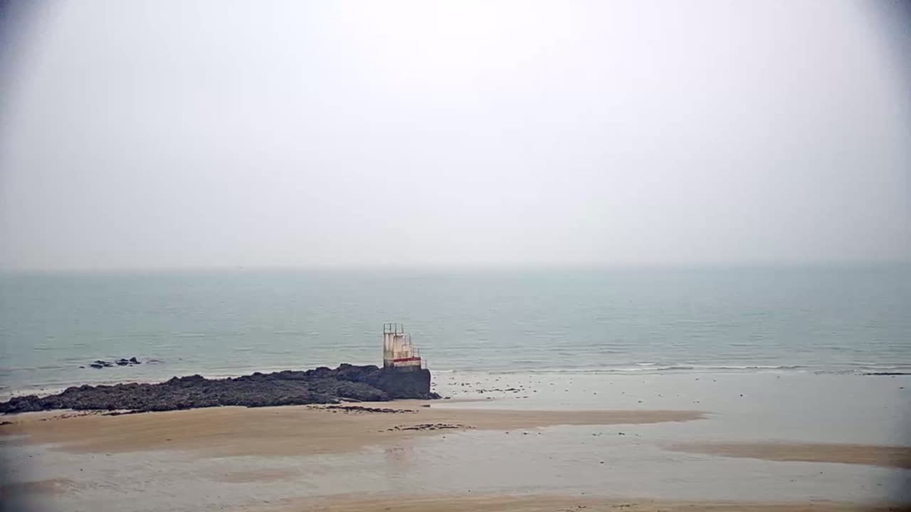 A hazy, overcast day reveals a calm ocean meeting a sandy beach where a weathered diving platform stands on dark rocks.