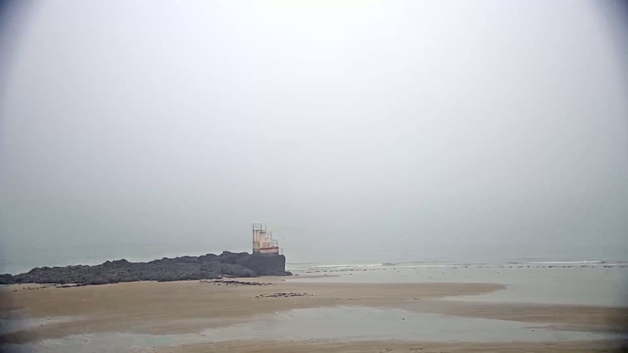A small, white structure sits on dark rocks near a calm, foggy ocean and a sandy beach.