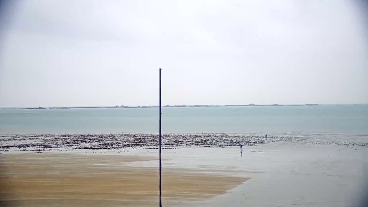 A sandy beach stretches toward a calm, light-blue ocean under an overcast sky, with a tall pole in the foreground and a few people visible in the distance.