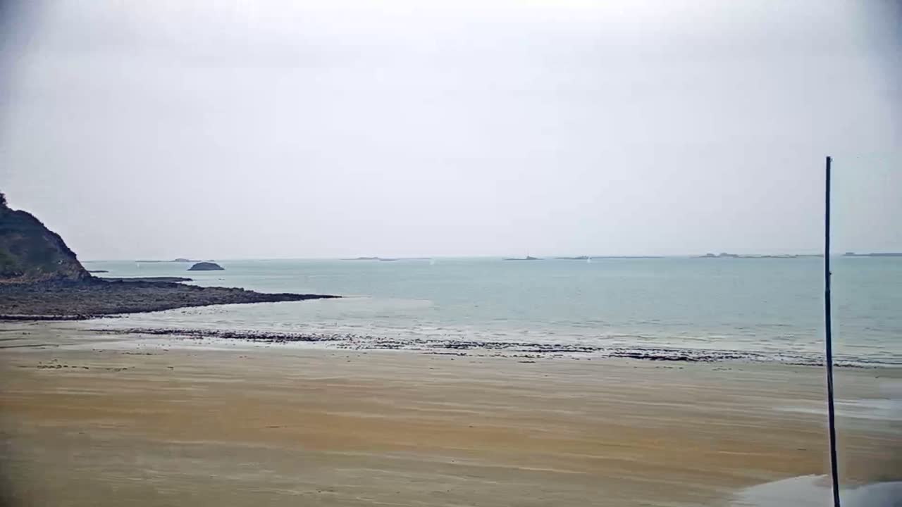 A sandy beach stretches towards a calm, light-grey sea under an overcast sky, with a dark cliff and several small islands visible in the distance.