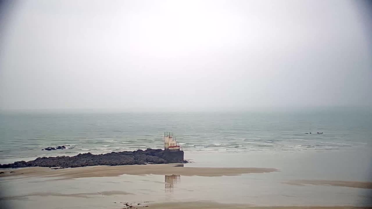 A small structure sits on dark rocks at the edge of a calm, foggy ocean, with a sandy beach in the foreground.