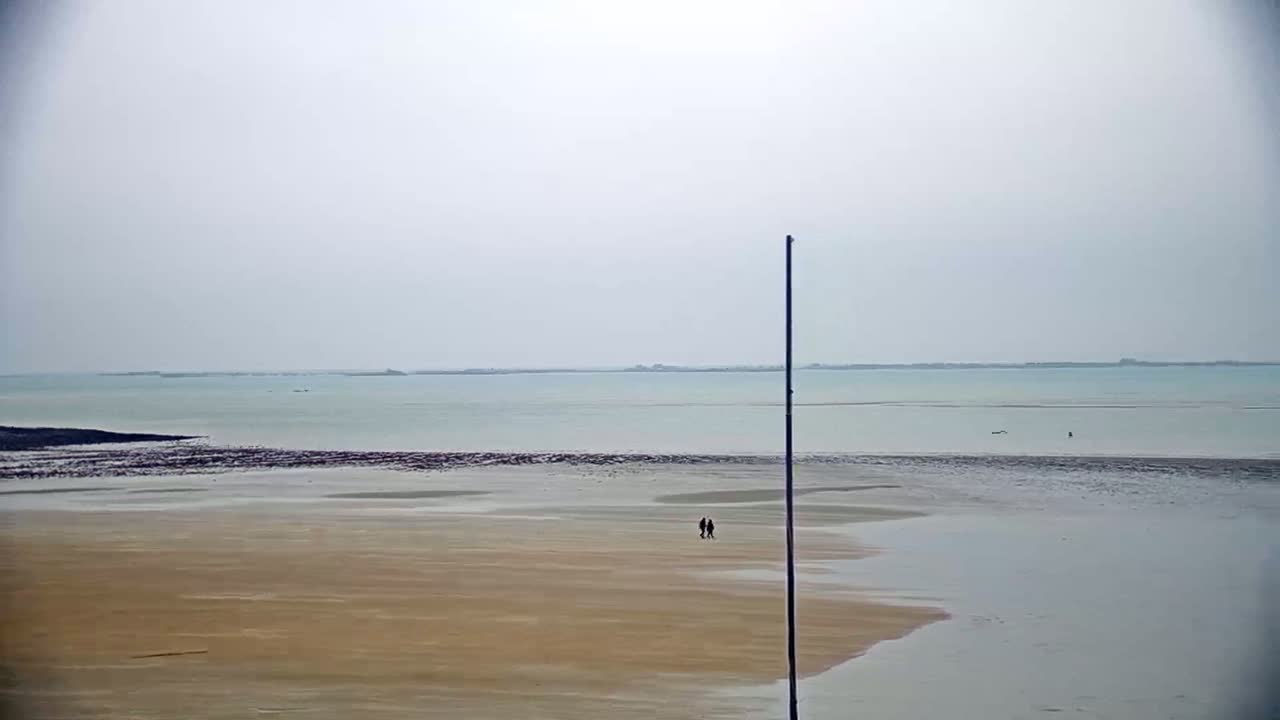 A wide, sandy beach stretches towards a calm, light-blue sea under an overcast sky, with two people walking in the distance.