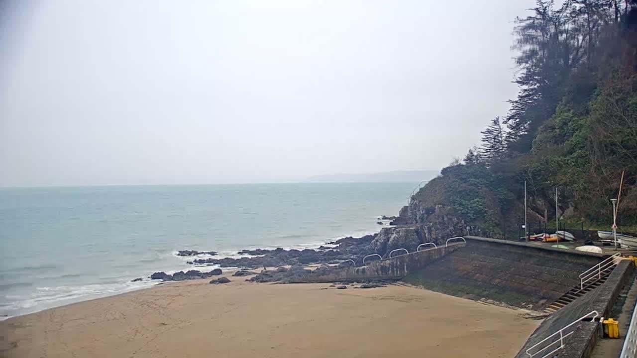 A sandy beach meets a calm, gray sea under an overcast sky, with rocky outcrops and a small boat launch area situated beside a verdant cliff.