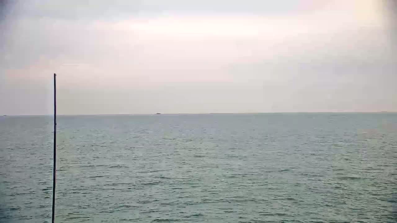 A tall, thin pole stands near a calm, grey sea under an overcast sky.