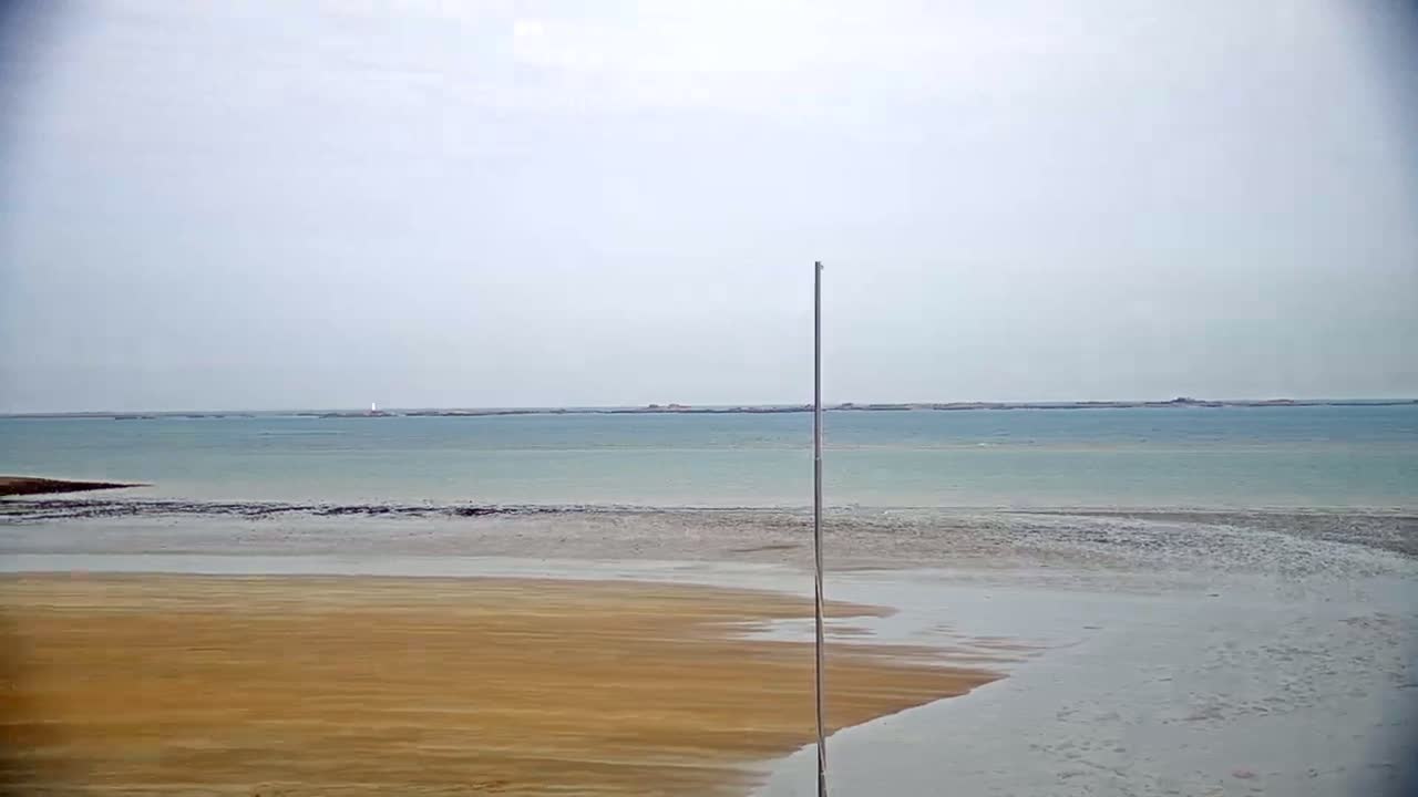 A tan sandy beach meets a calm, light-blue sea under a cloudy sky, with a tall pole standing near the shore.