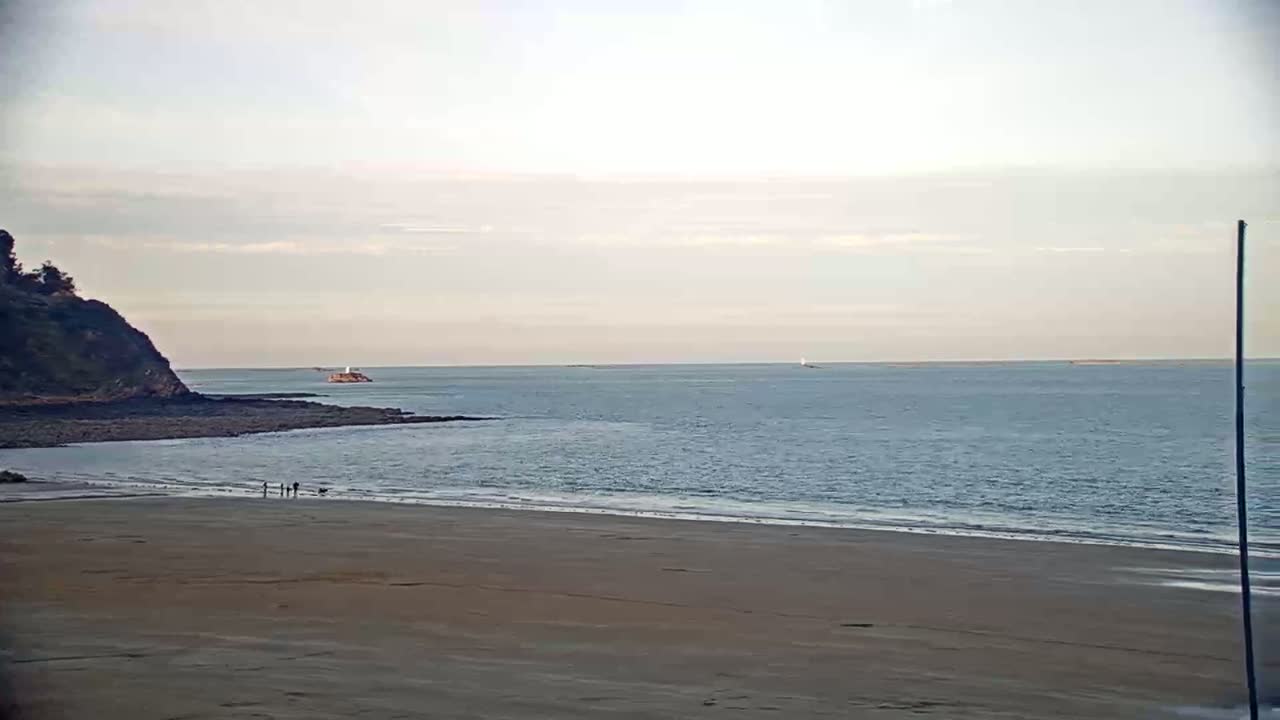 A sandy beach stretches out to a calm ocean under a pale sky, with a small group of people walking near the water's edge and a distant rocky outcrop.