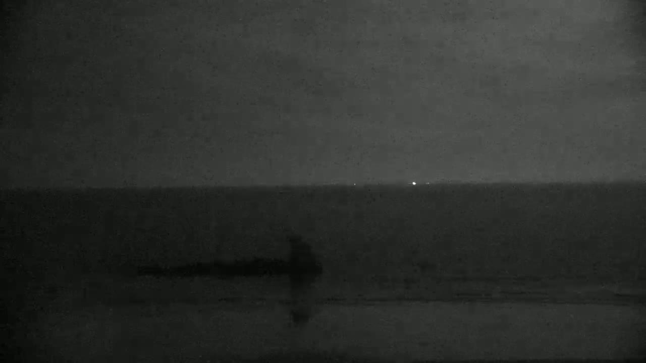 A dark, nighttime beach scene shows a lone figure standing near the water's edge under a mostly cloudy sky.