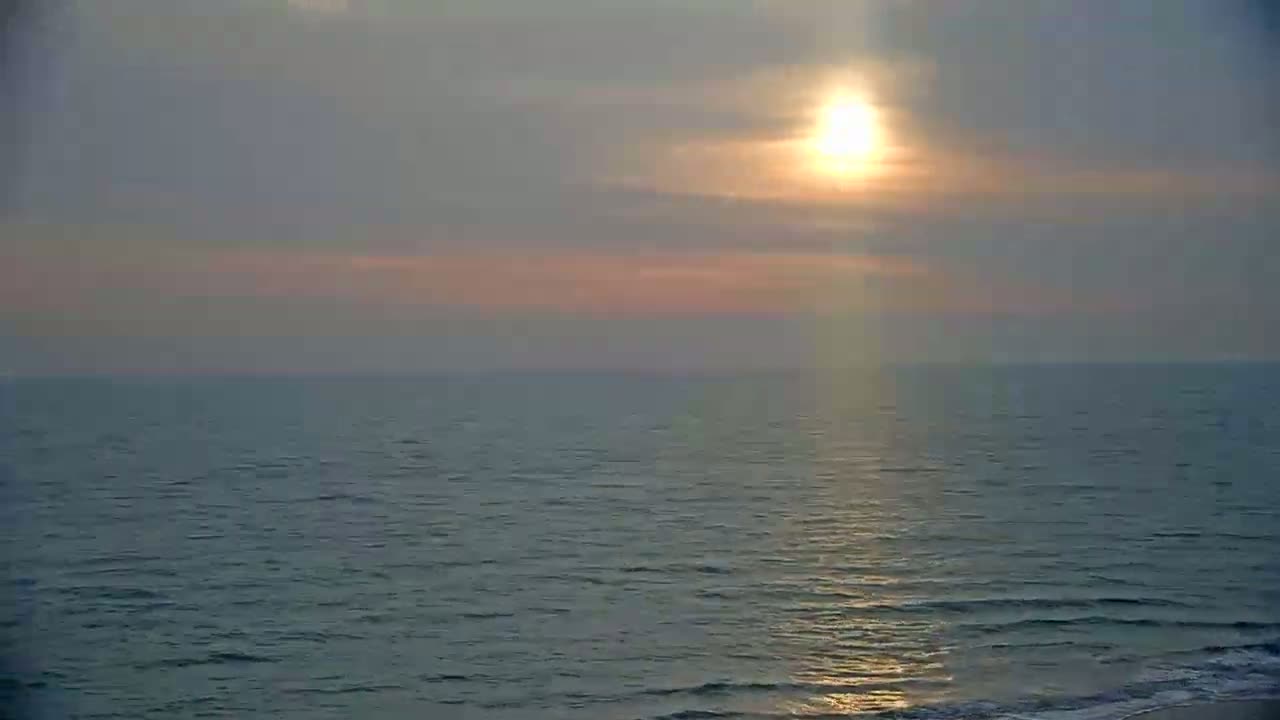 A calm ocean with small waves is seen at sunset under a mostly cloudy sky.