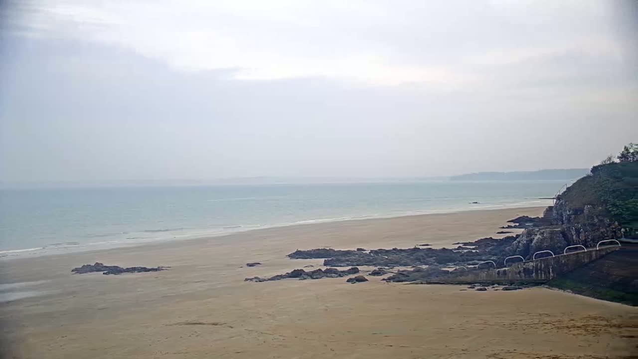 A wide sandy beach stretches to a calm, light blue ocean under an overcast sky, with dark rocks near the shoreline and a green cliff in the background.