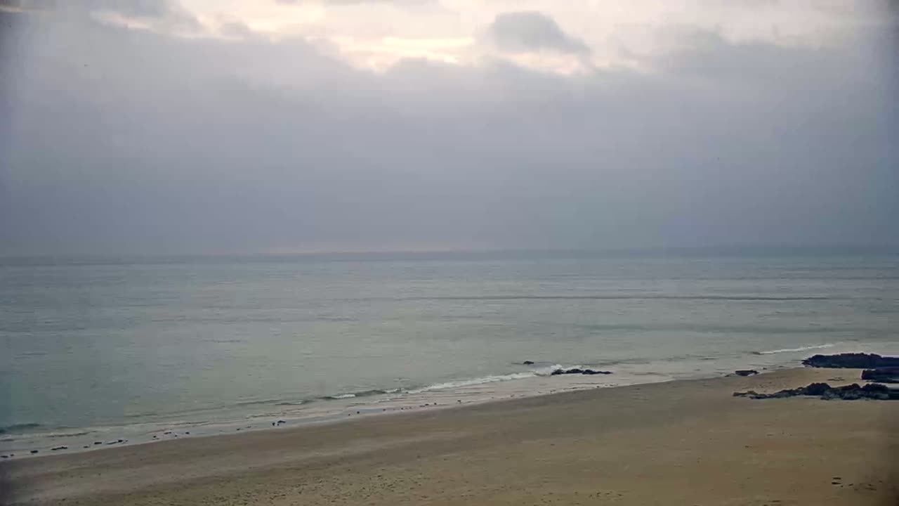 A sandy beach meets a calm, light-grey sea under a cloudy sky.