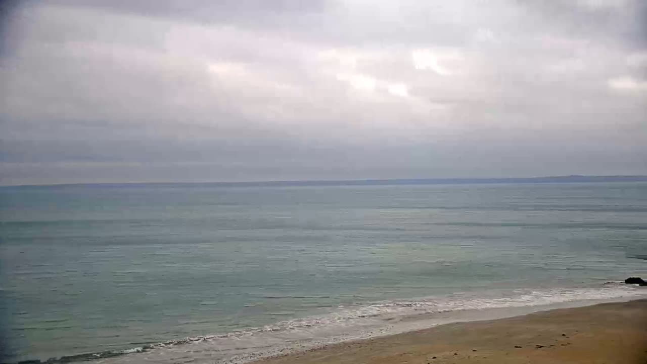 A sandy beach meets a calm, light-green ocean under a mostly cloudy sky.