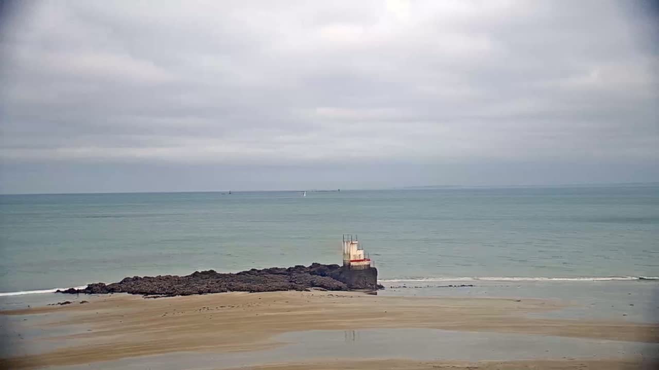 A small, white structure sits atop dark rocks on a sandy beach, with a calm, gray-sky ocean in the background.
