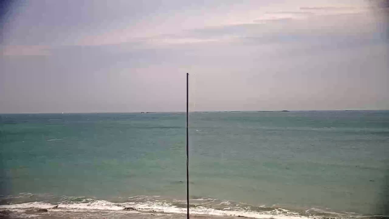 A tall, thin pole stands in front of a calm, teal ocean meeting a sandy beach under a mostly cloudy sky.