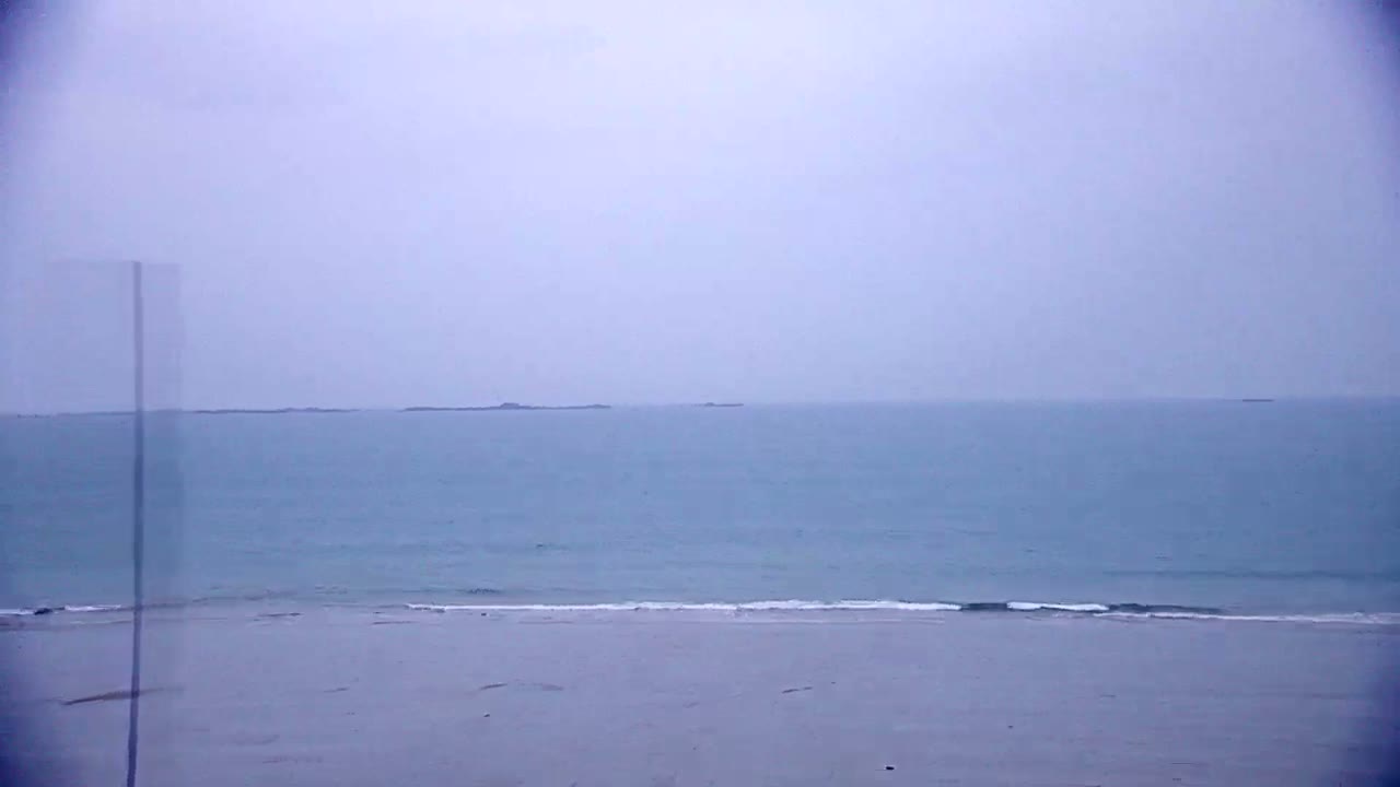 A calm, light-blue ocean meets a pale sandy beach under an overcast sky.