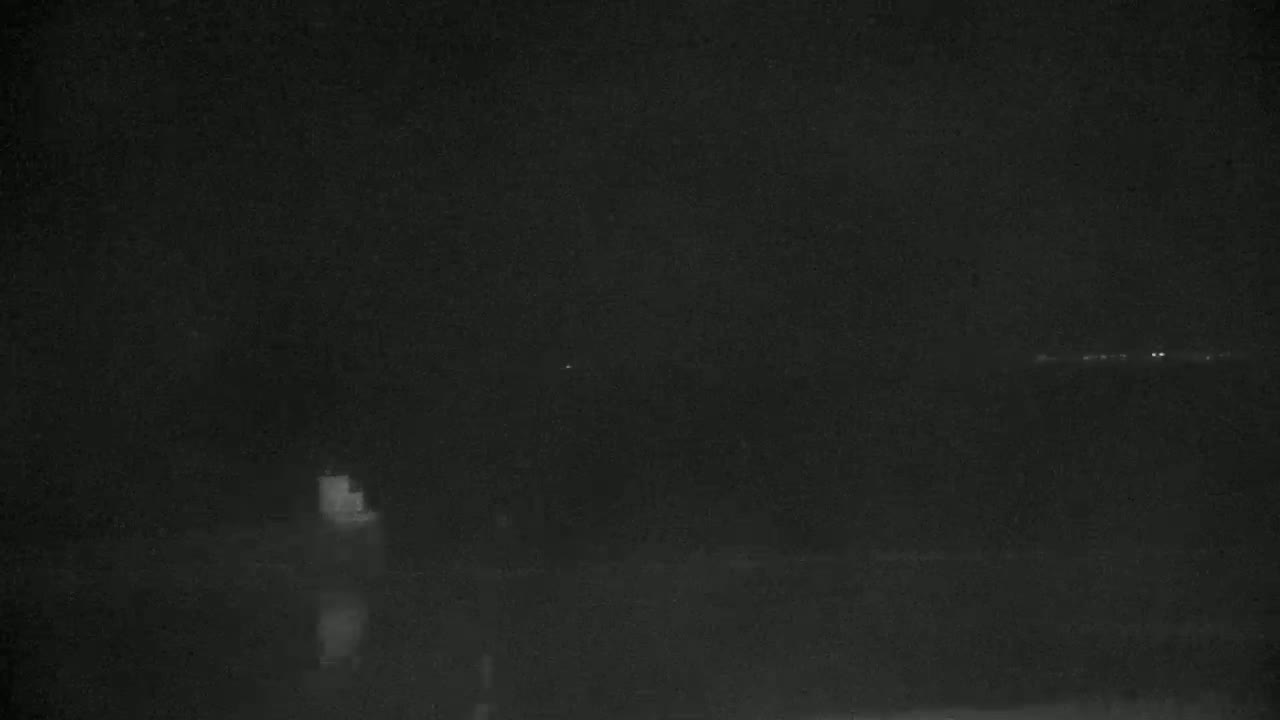 A dark, nighttime scene shows a body of water with a faintly illuminated structure on the left and a distant, faint light source on the right.