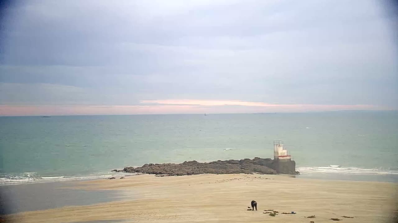 A person is standing on a sandy beach near a rocky outcrop and a small structure, under a cloudy sky with a calm, light-green sea.