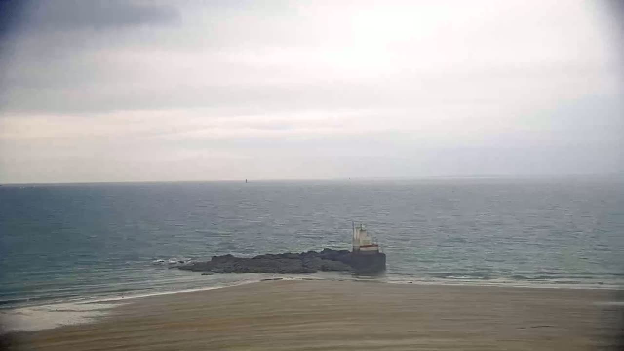 A small structure sits on rocks near a sandy beach and a calm, gray sea under a cloudy sky.