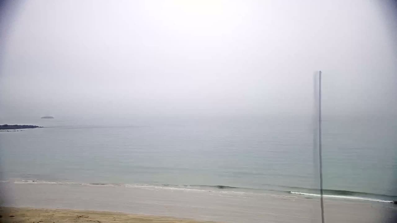 A hazy, foggy day reveals a calm ocean meeting a sandy beach, with a small island visible in the distance.