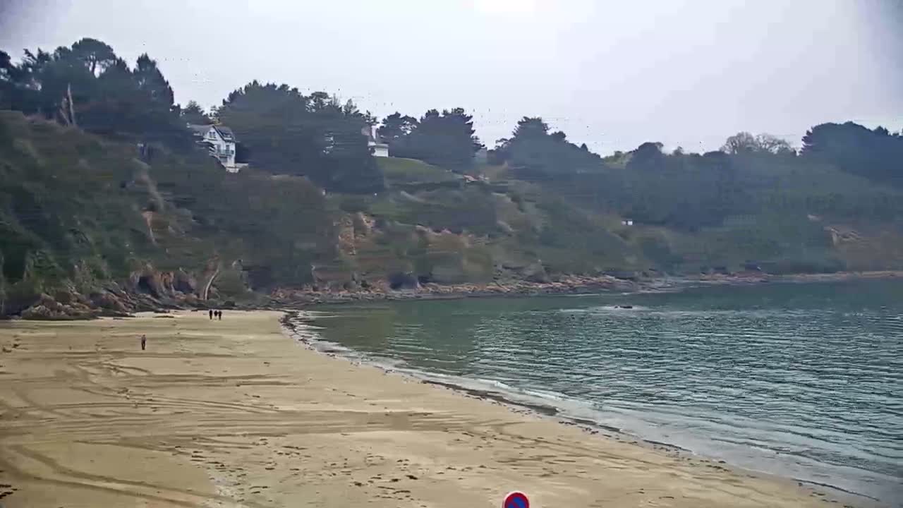 A sandy beach curves gently into calm, gray-green water, backed by a verdant, rocky hillside dotted with trees under an overcast sky.