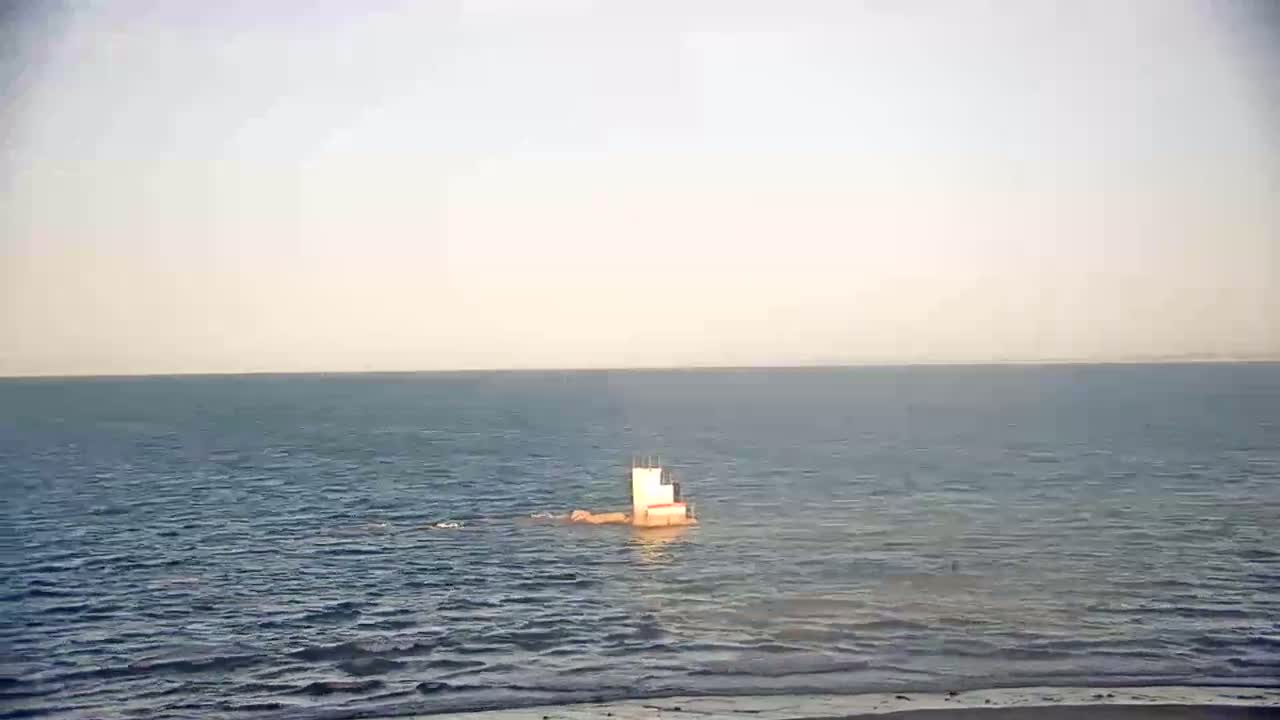 A small, light-colored structure sits in calm, blue water under a hazy sky.