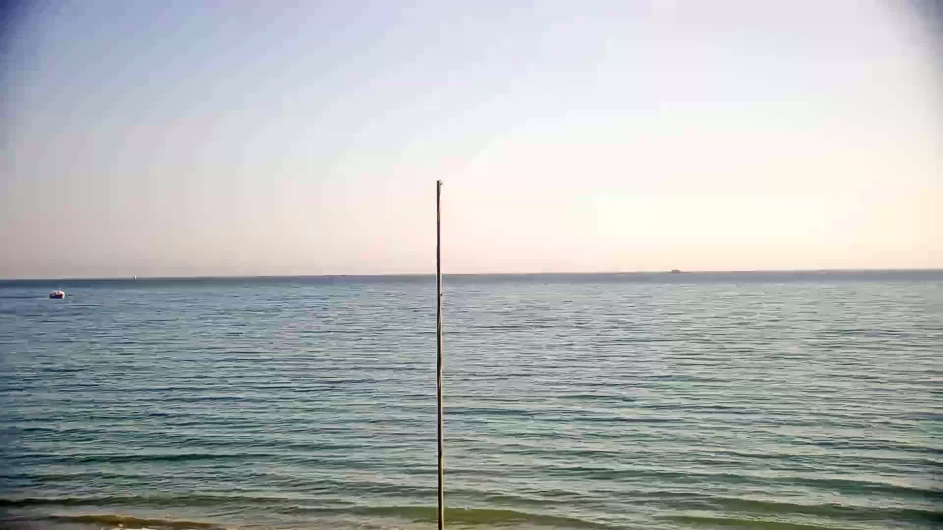 A tall, thin pole stands near a calm, blue-green ocean under a clear, light-blue sky.