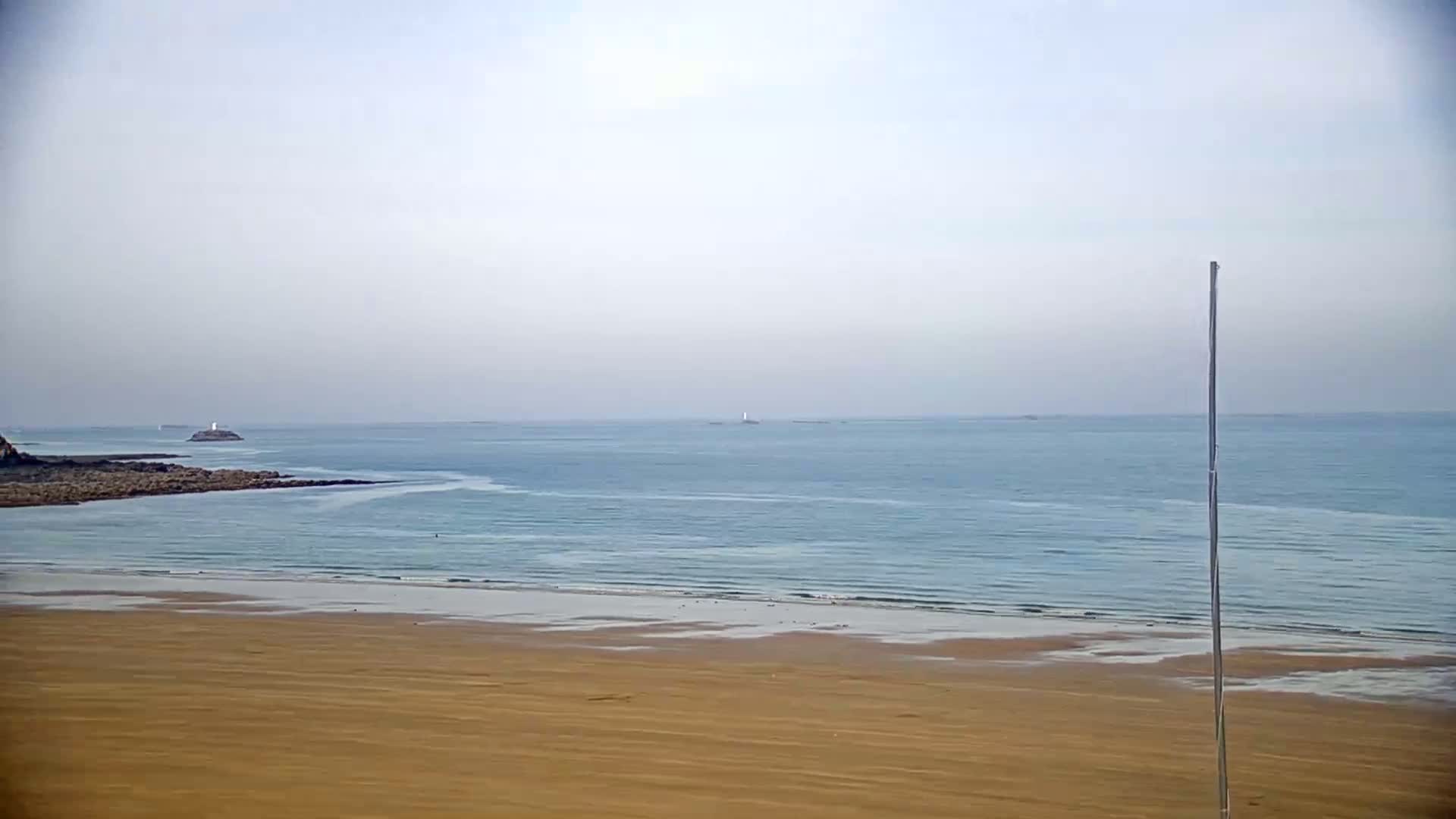 A long sandy beach stretches to a calm, light-blue ocean under a hazy sky, with a tall pole standing near the lower right.