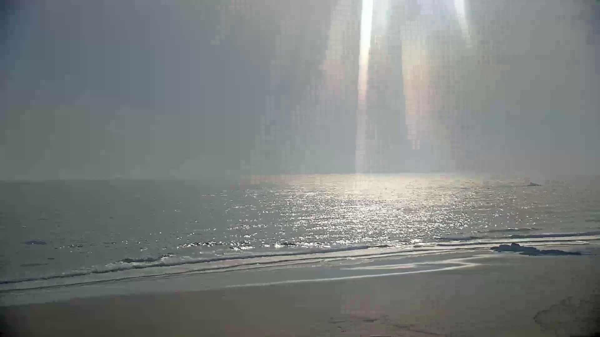 A hazy sun shines down on a calm ocean meeting a sandy beach.