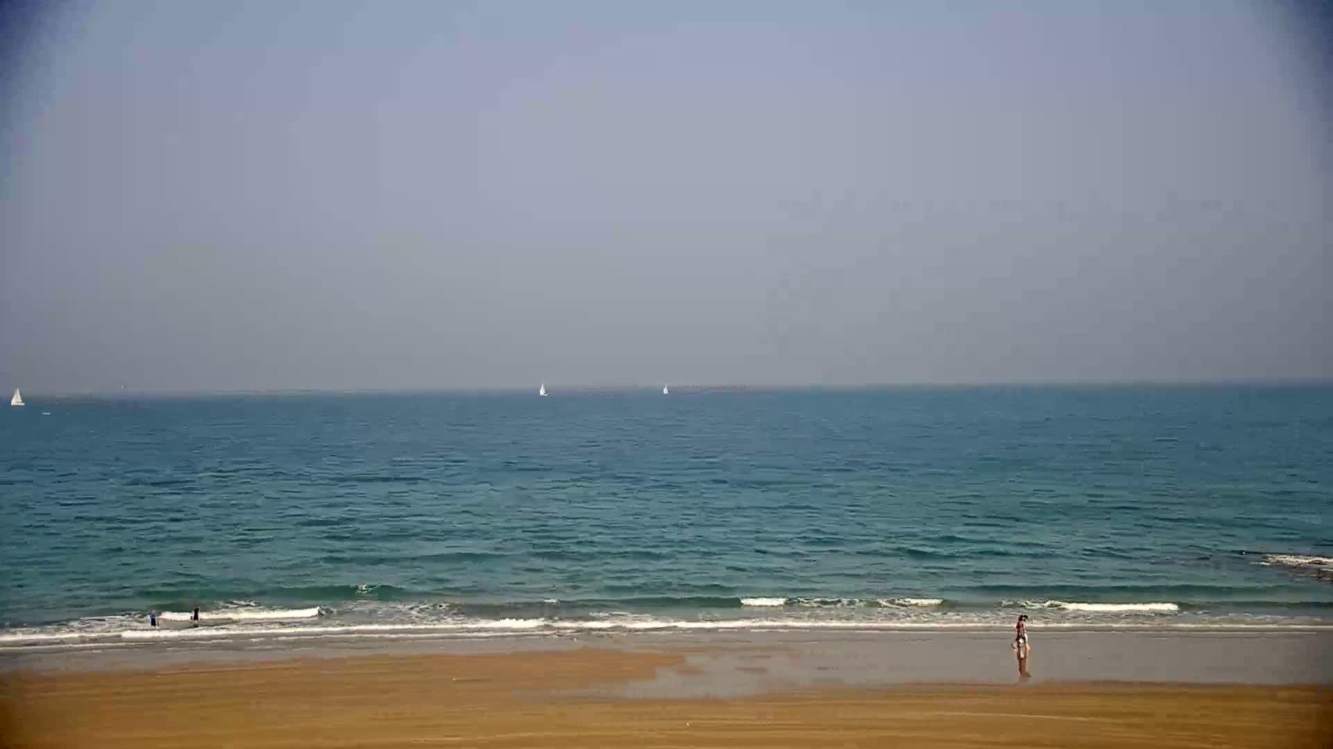 A calm, hazy ocean with a few sailboats on the horizon meets a wide, sandy beach where a few people are standing near the water's edge.