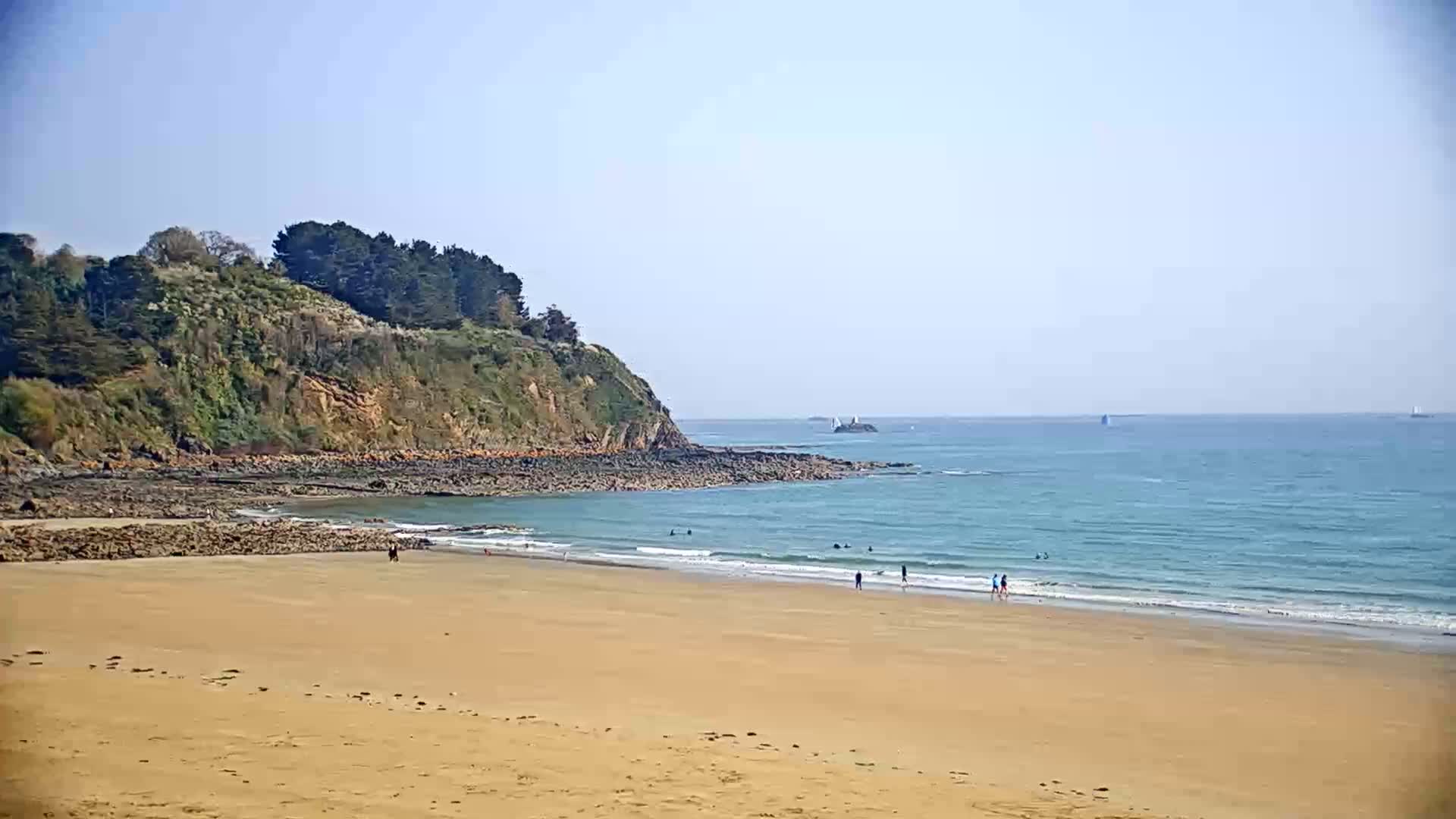 A sandy beach stretches out to a calm, light blue ocean meeting a rocky shoreline topped with green vegetation under a clear, bright sky.