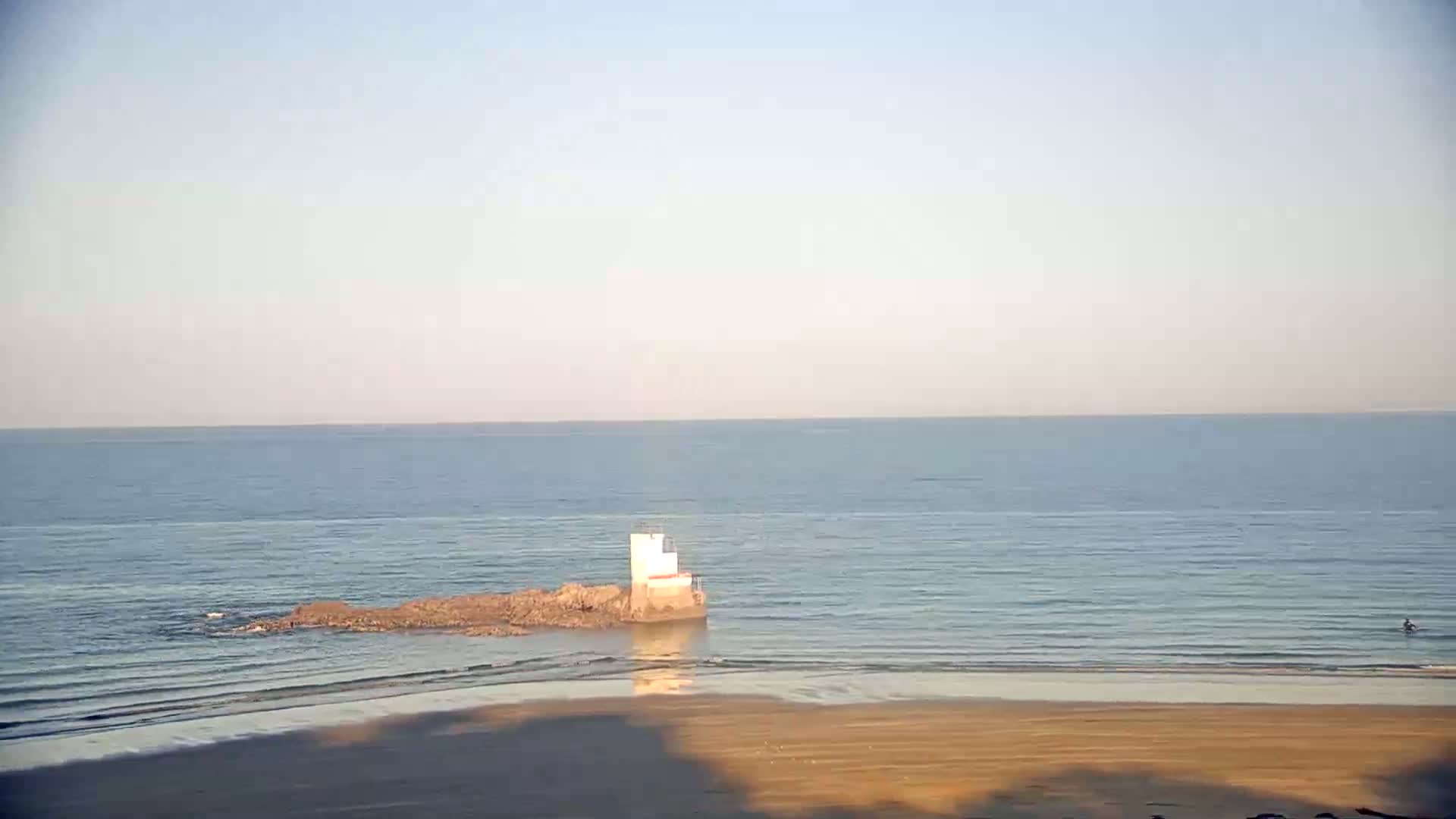 A small, white structure sits on rocks jutting out from a sandy beach into a calm, blue ocean under a clear sky.