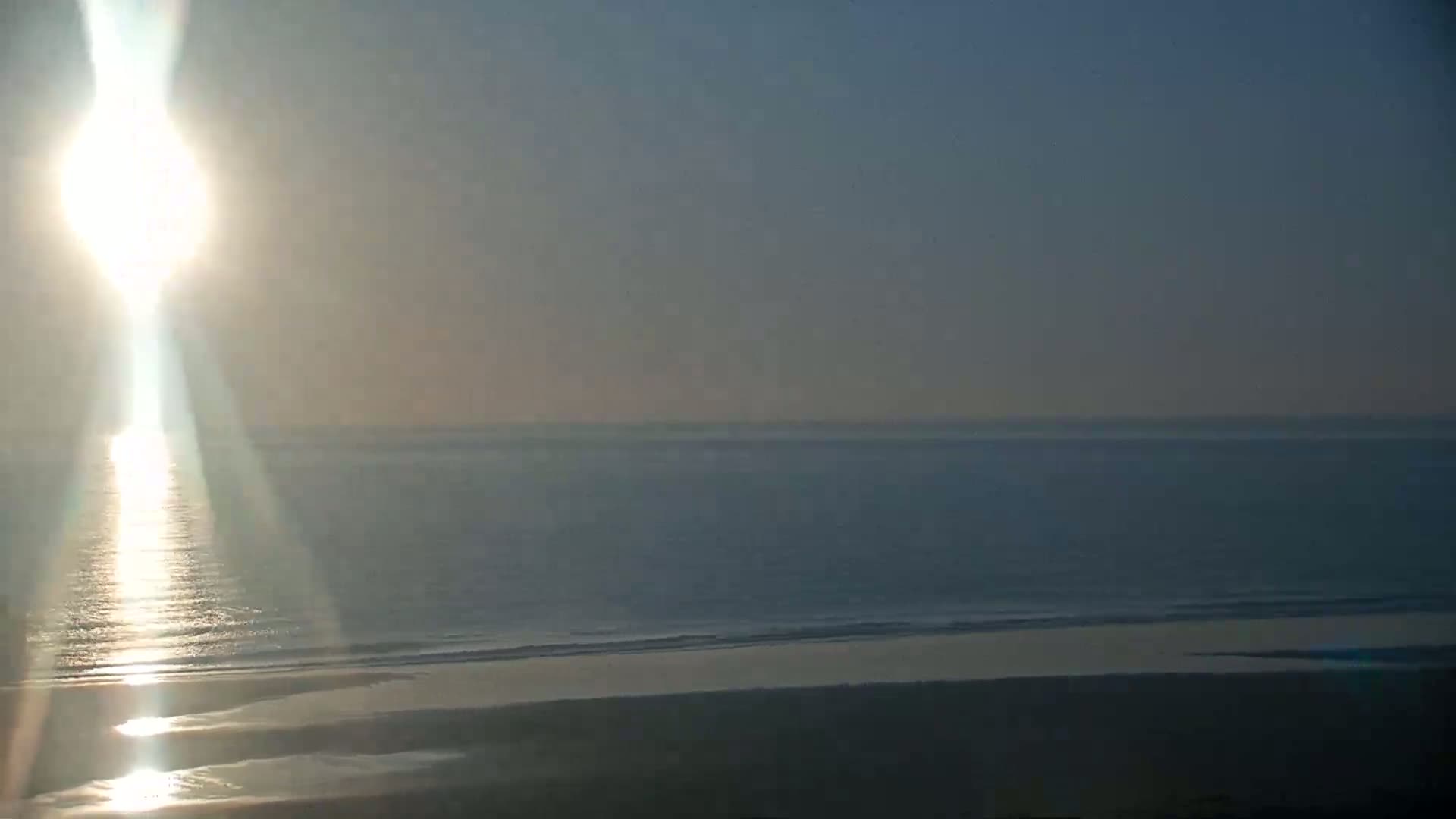 A bright sun shines over a calm ocean and sandy beach.