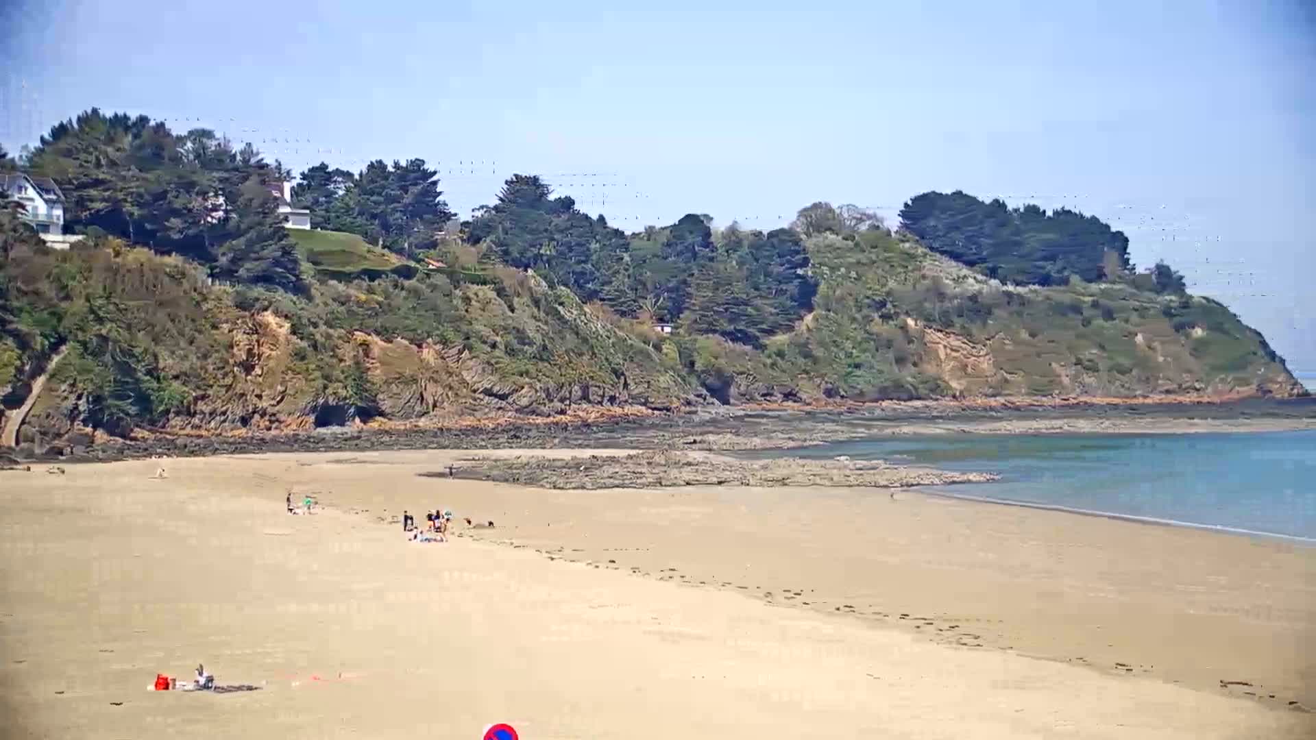 A sandy beach stretches out to calm blue water, bordered by a rocky, tree-covered headland under a clear blue sky.