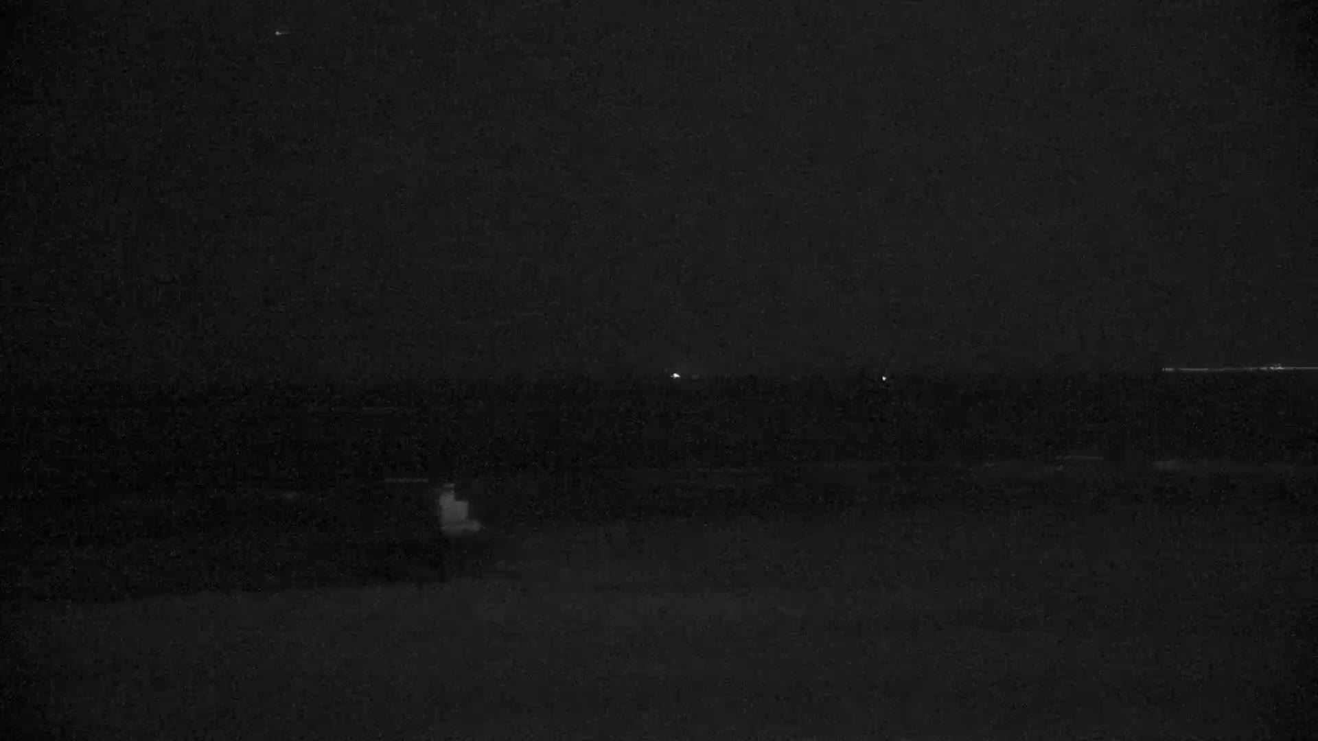 A dark, nighttime scene shows a flat, dark landscape with a few faint lights in the distance under a clear, dark sky.