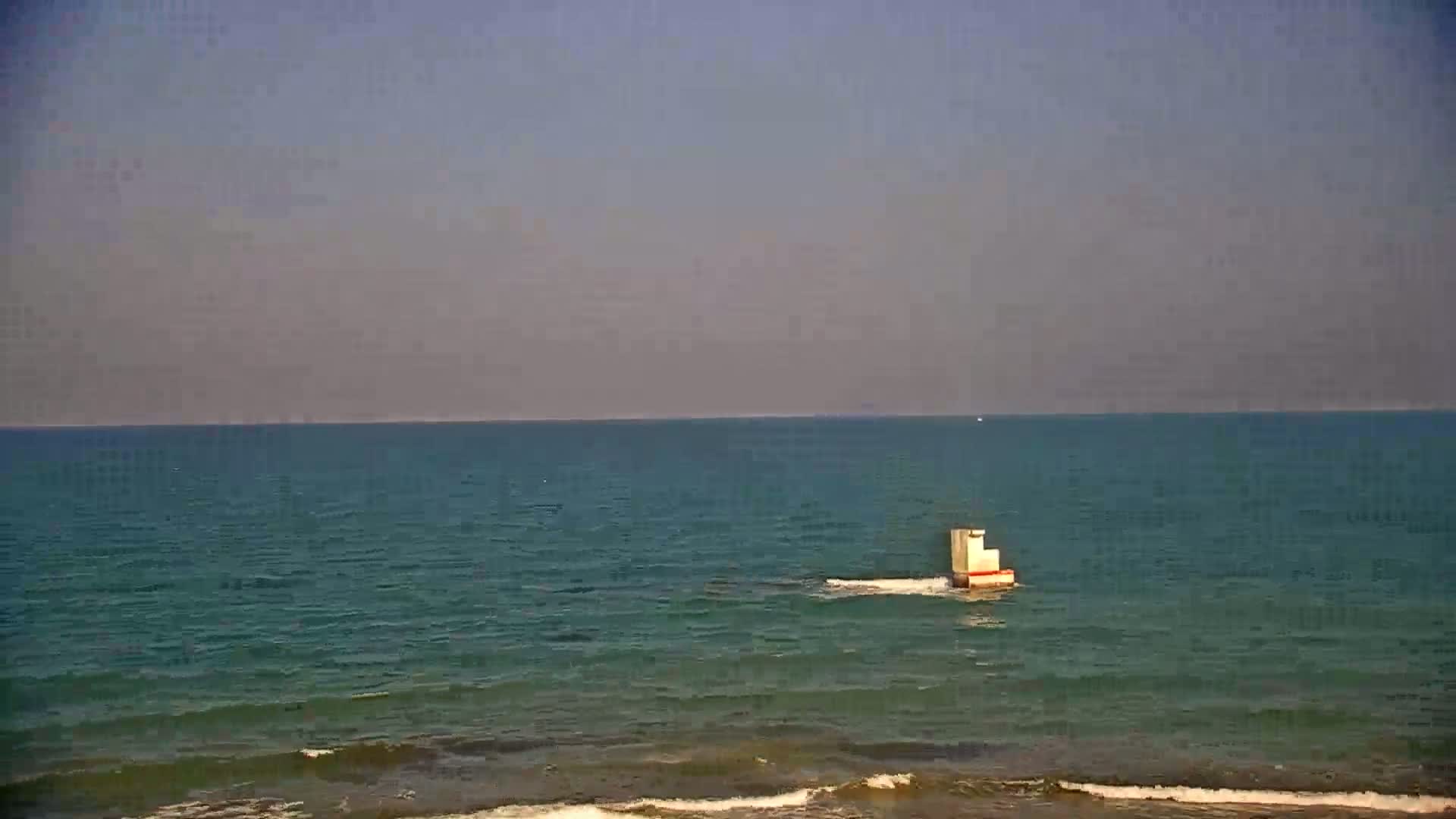 A small, light-colored structure sits in a calm, teal sea under a hazy sky.