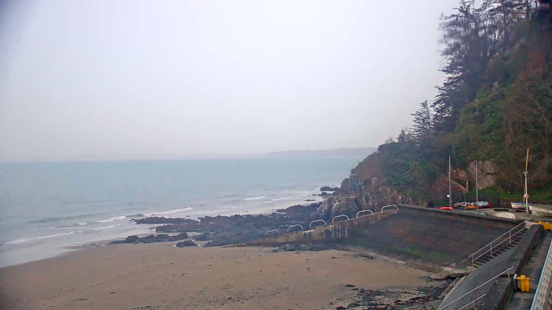 A sandy beach stretches to a calm, gray sea under an overcast sky, with a rocky shoreline and a wooded cliff in the background.