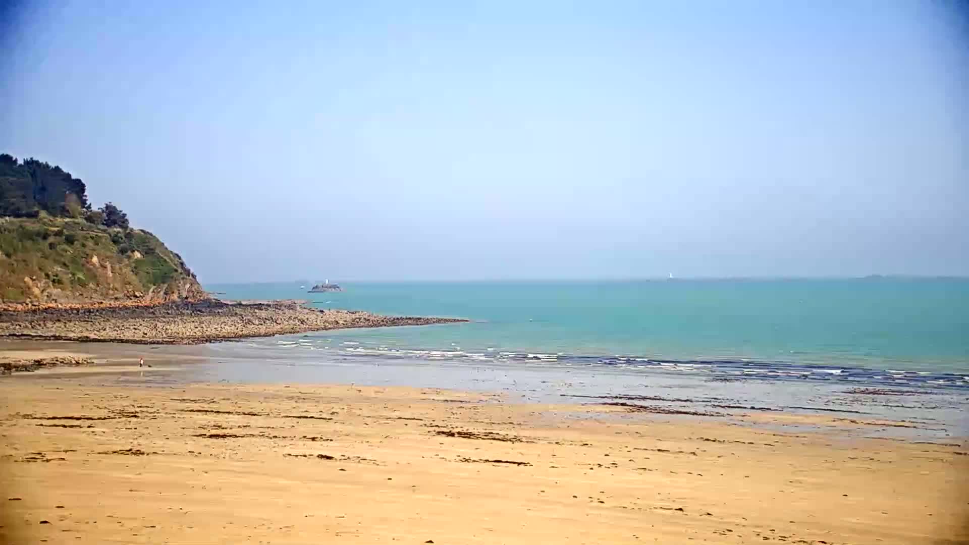 A sandy beach stretches to a calm, teal ocean under a clear blue sky, backed by a rocky, green-covered headland.