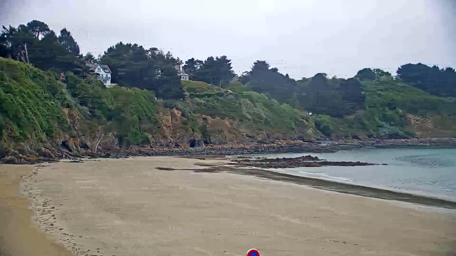 A sandy beach curves gently around a rocky cove, backed by a verdant cliff with houses perched on top, under an overcast sky.