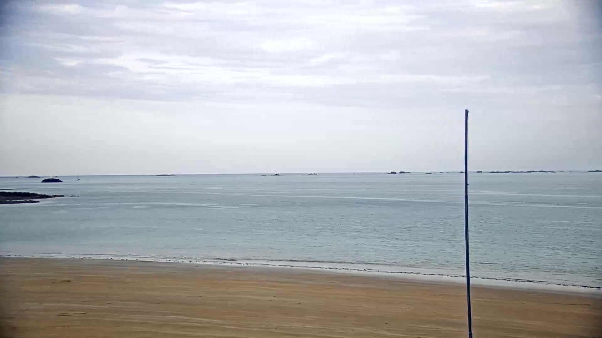 A sandy beach stretches to a calm, light-blue ocean under a cloudy sky, with a tall pole standing near the shore.