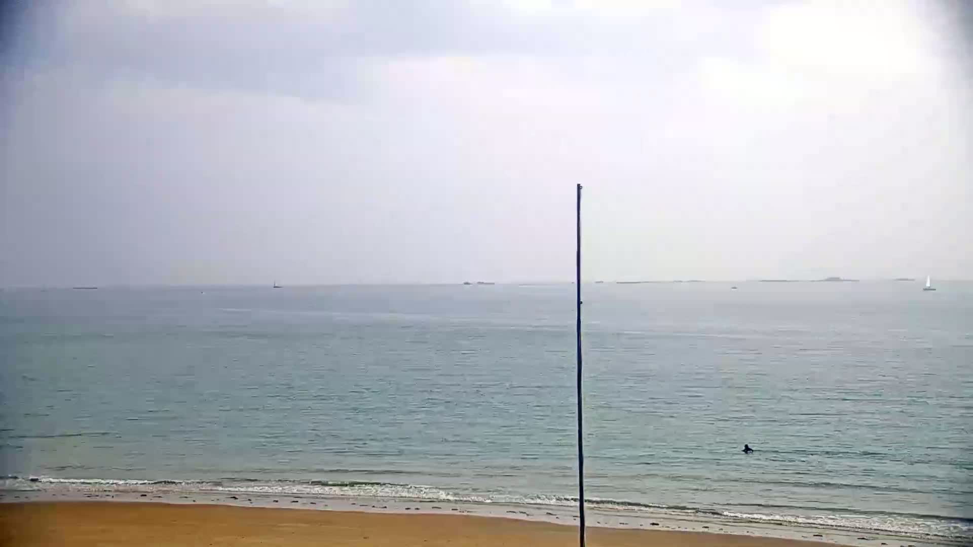 A sandy beach meets a calm, light-blue ocean under a hazy sky, with a single person visible in the water and a few distant boats.