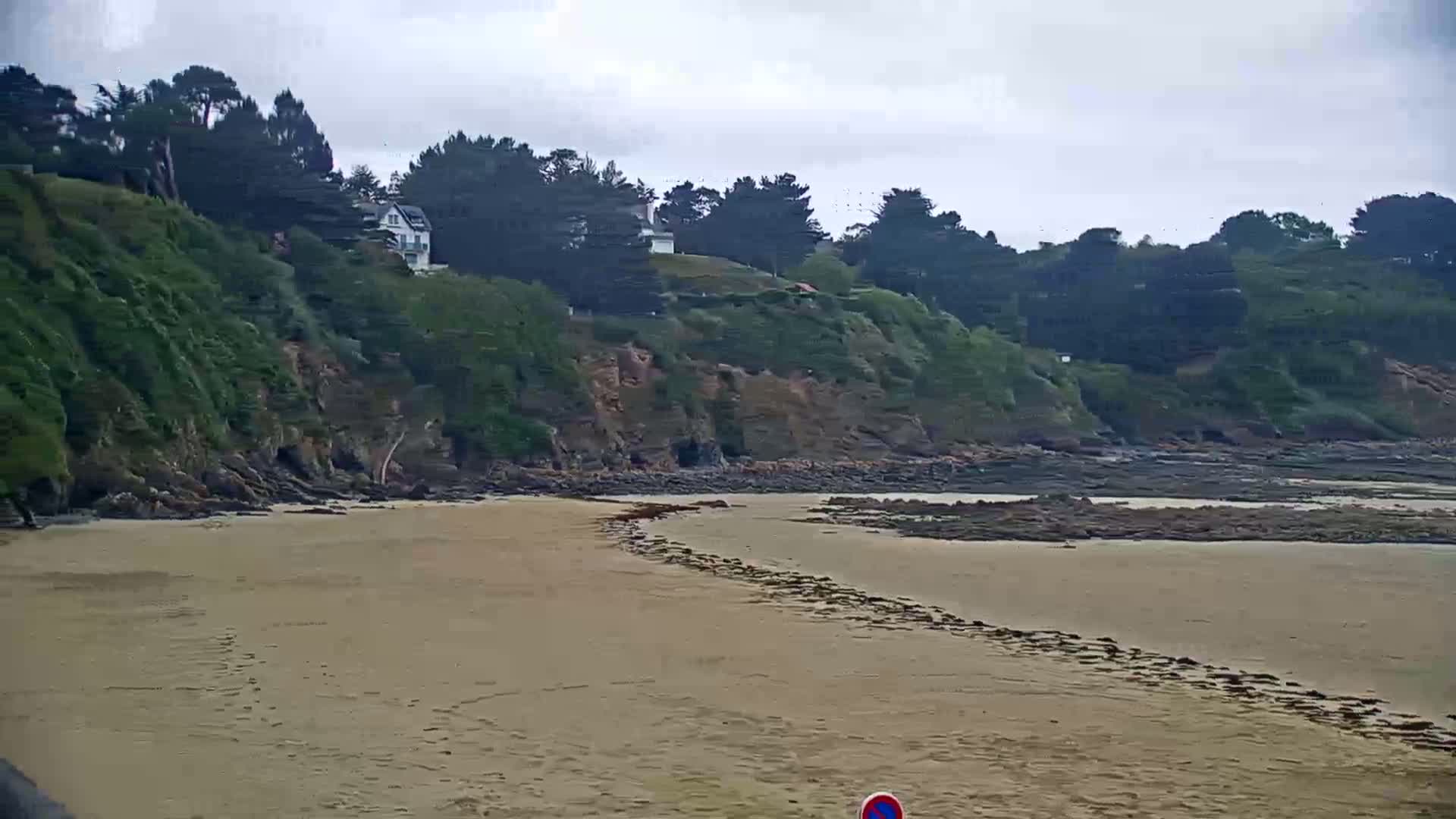 A sandy beach stretches out to a rocky shoreline under an overcast sky, backed by a verdant cliff with houses nestled amongst the trees.