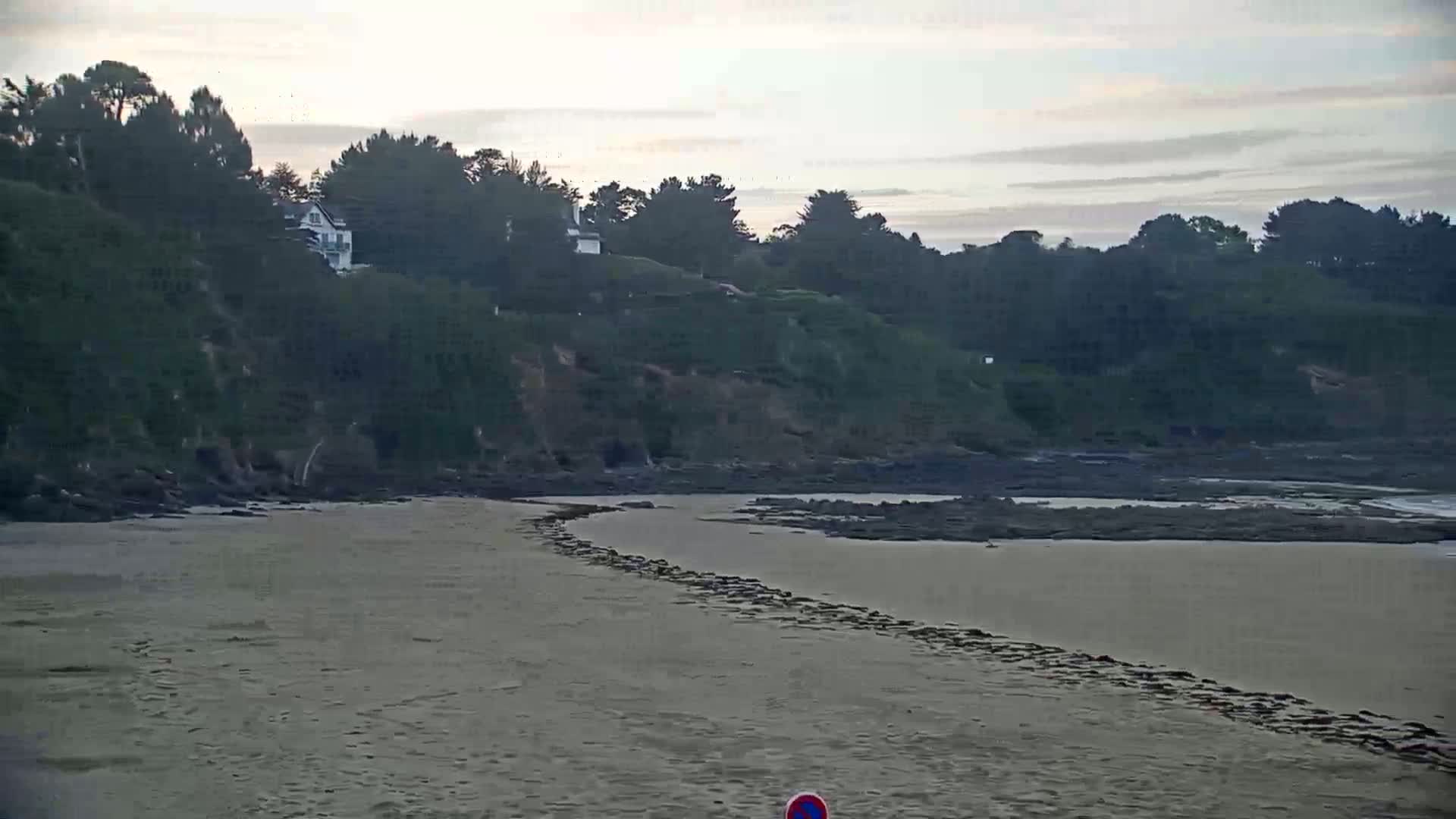 A sandy beach curves around a calm, shallow body of water, bordered by a rocky shoreline and a hillside covered in dark green trees and a few white houses under an overcast sky.