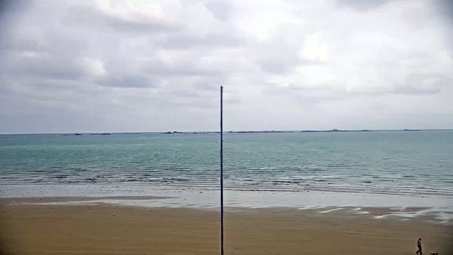 A sandy beach stretches to a calm, teal sea under a cloudy sky, with a tall pole standing in the middle of the frame and a lone person walking a dog in the distance.