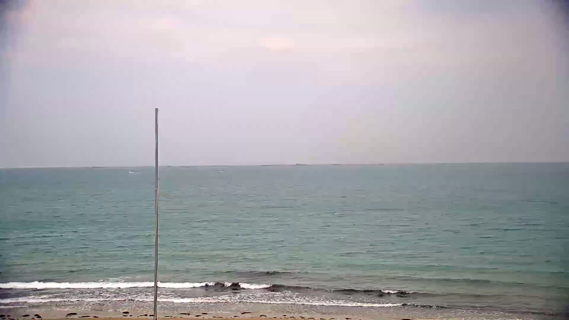 A mostly cloudy day shows a calm sea meeting a sandy beach with small waves gently breaking.