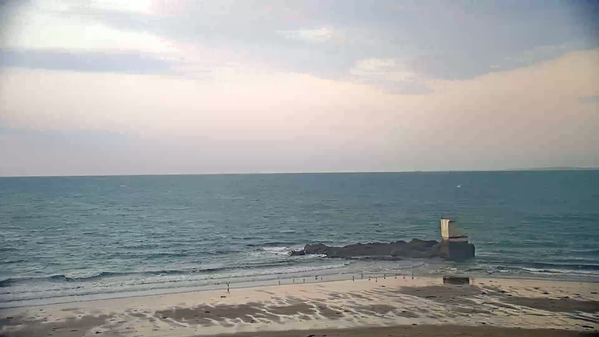 A sandy beach with a few people on it stretches to a calm, blue ocean meeting a rocky outcrop with a small, light-colored structure on top, under a mostly cloudy sky.