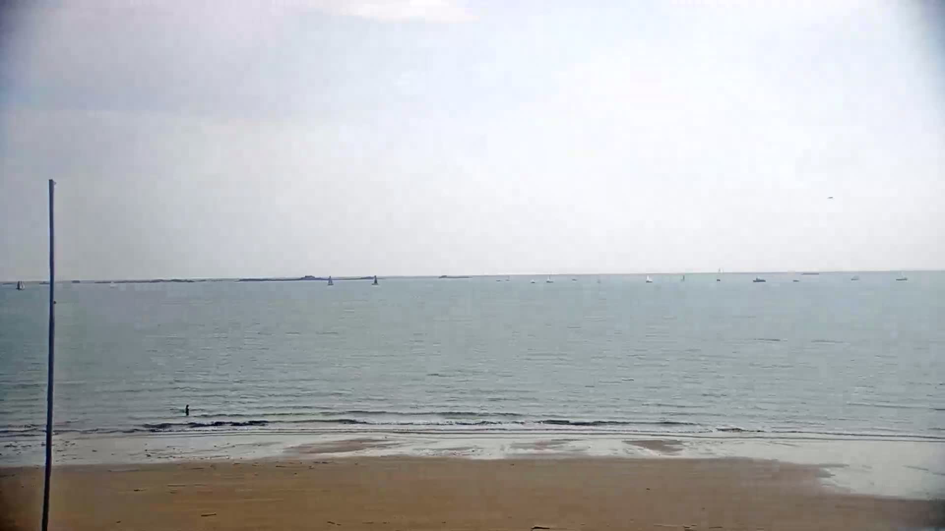 A calm, overcast day at a beach shows a flat expanse of water with several small boats in the distance and a sandy shore in the foreground.
