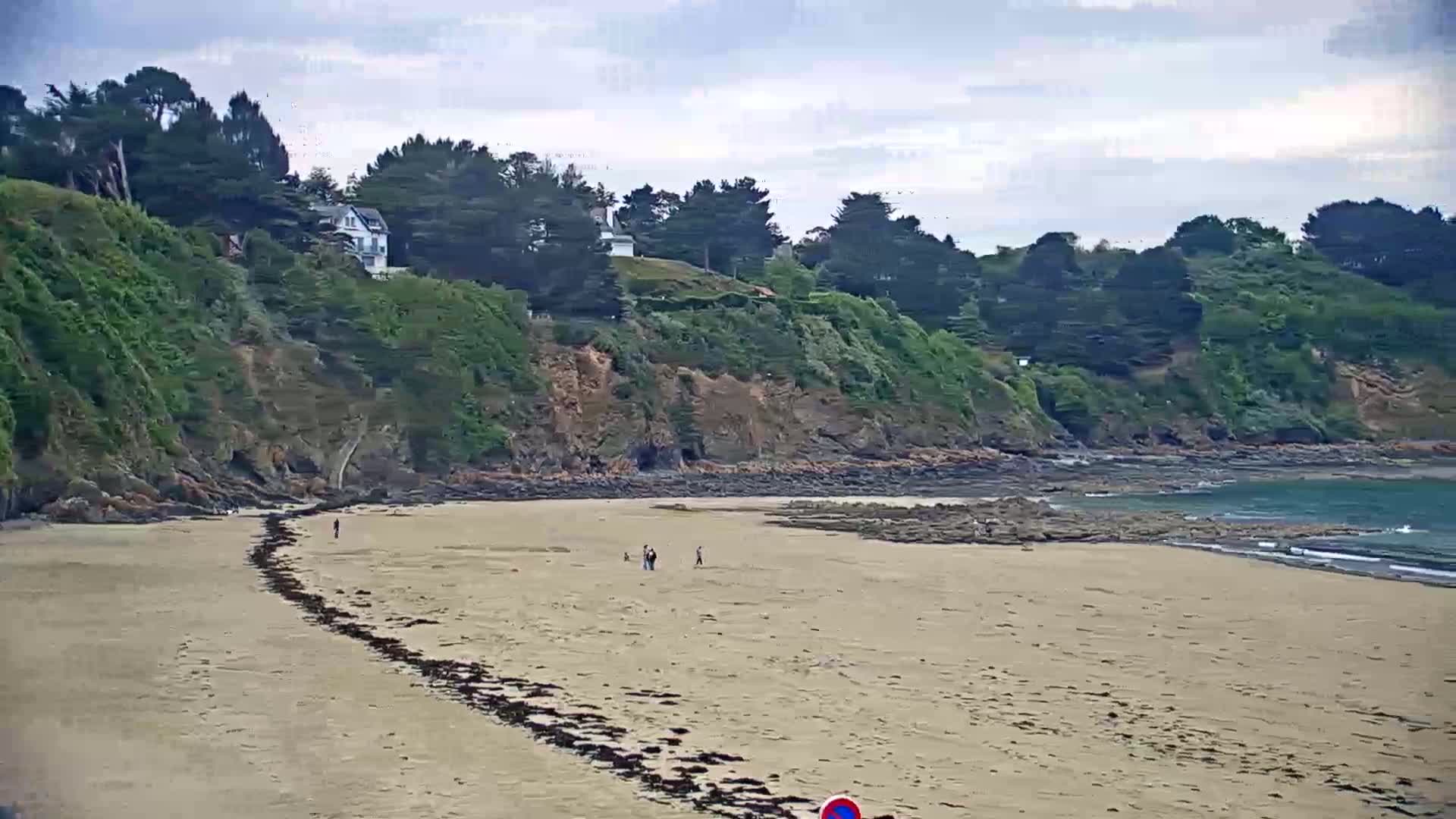 A sandy beach stretches out to a calm, teal ocean under a cloudy sky, backed by a rocky, green-covered cliff with several houses visible atop it.