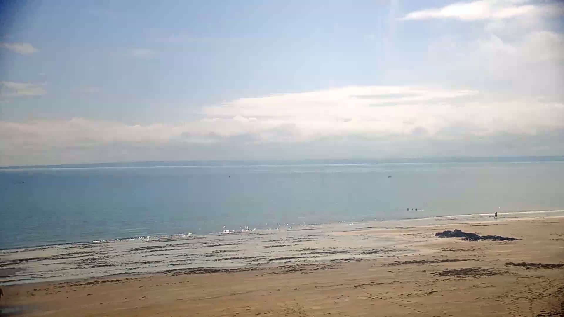 A sandy beach stretches to a calm, light blue sea under a mostly clear, bright sky with some scattered clouds.